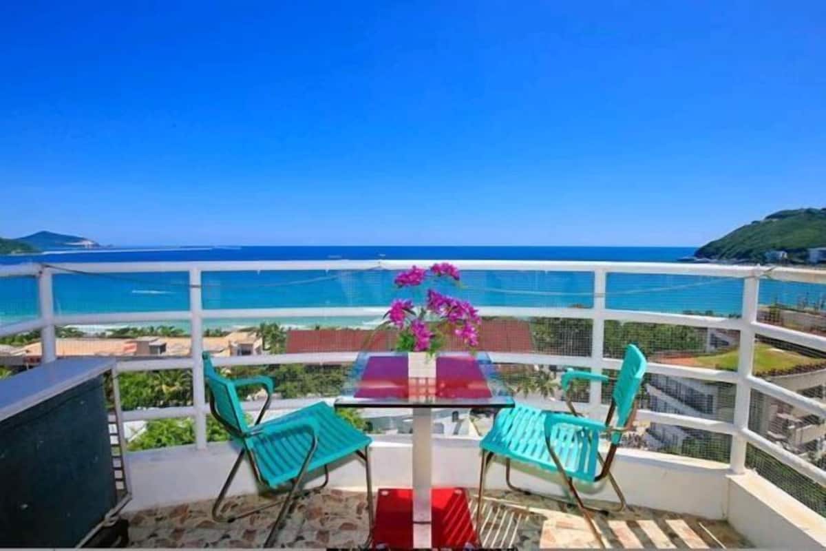 Een balkon met een glazen tafel en stoelen, met uitzicht op een strand en de oceaan.