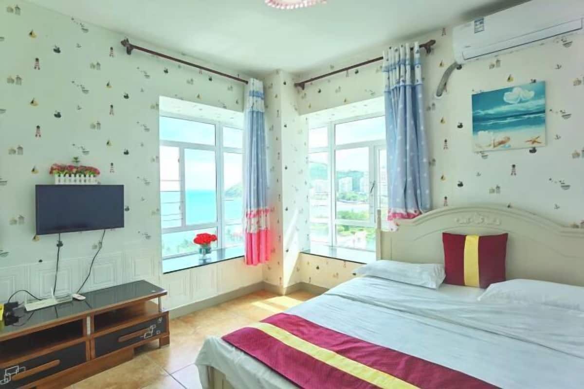 Een slaapkamer met een bed, een televisie, een houten kast en grote ramen met gordijnen.