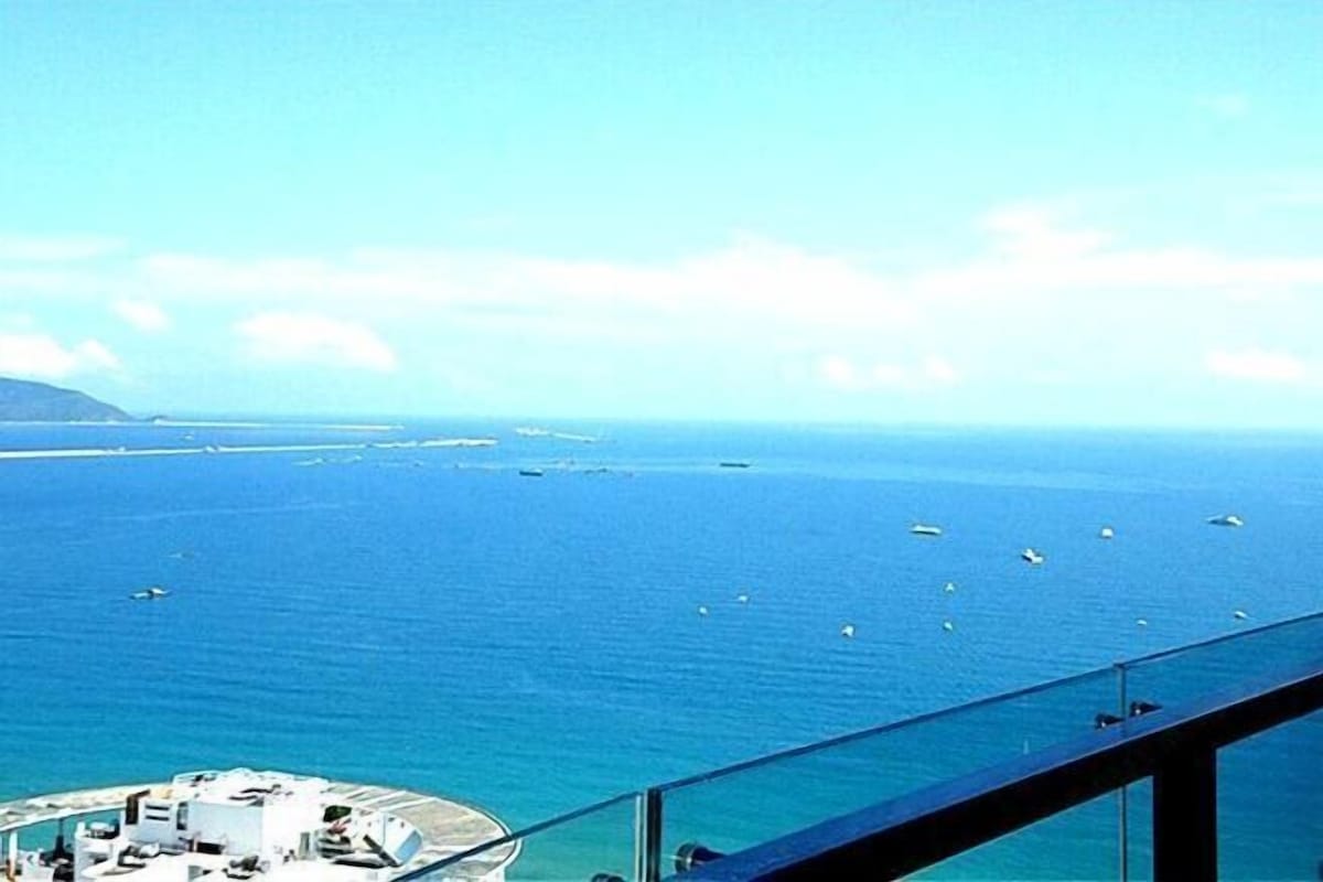 Vista desde un balcón sobre un mar tranquilo con barcos, una costa distante y un cielo despejado.