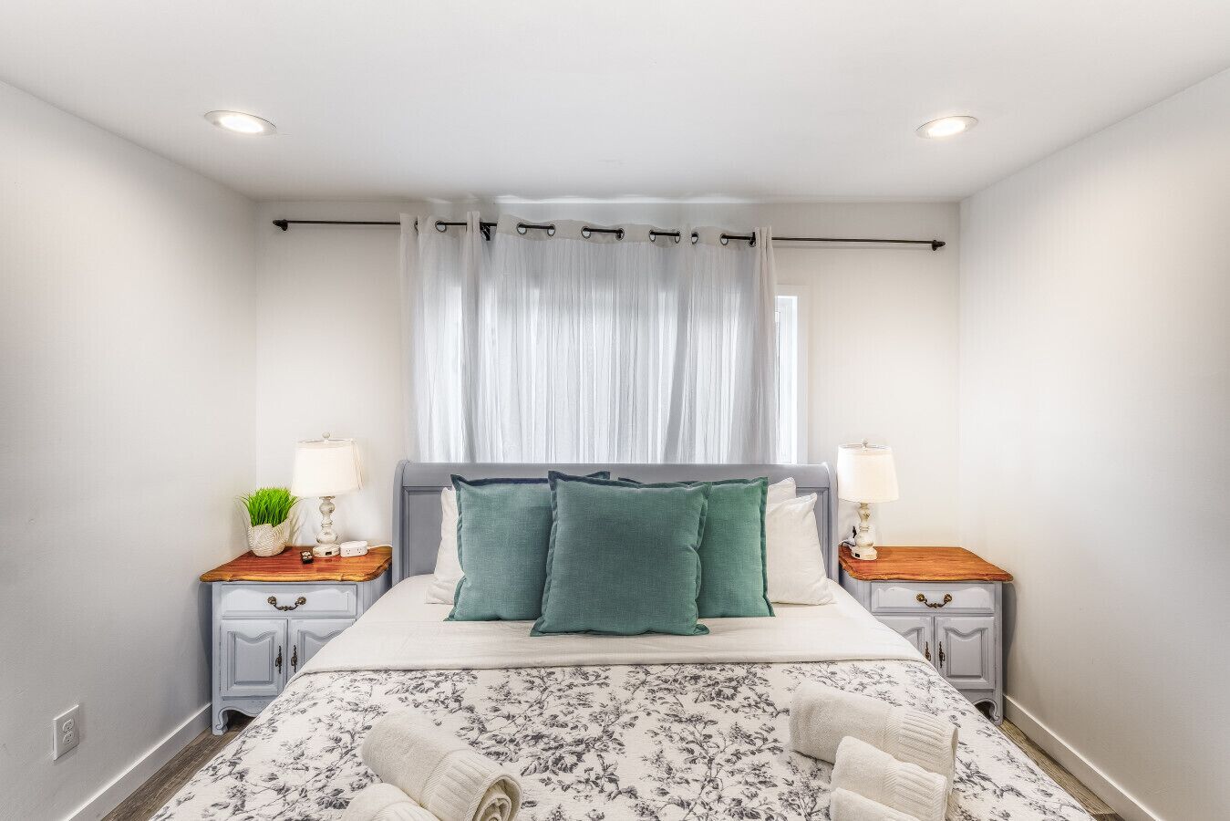 Quarto bem arrumado com cama, duas mesas de cabeceira, uma luminária e uma janela com cortinas finas, além de uma colcha com padrão floral.