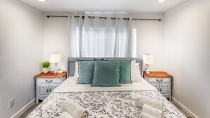 Quarto bem arrumado com cama, duas mesas de cabeceira, uma luminária e uma janela com cortinas finas, além de uma colcha com padrão floral.