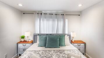 Quarto bem arrumado com cama, duas mesas de cabeceira, uma luminária e uma janela com cortinas finas, além de uma colcha com padrão floral.