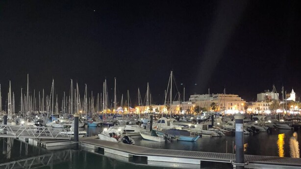 Ein Yachthafen bei Nacht mit vertäuten Booten, einer Brücke und Gebäuden im Hintergrund.