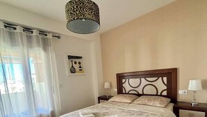 Ein Schlafzimmer mit Bett, zwei Nachttischen, einer Pendelleuchte, einem Fenster mit durchsichtigen Vorhängen und einem an der Wand hängenden Kunstwerk.