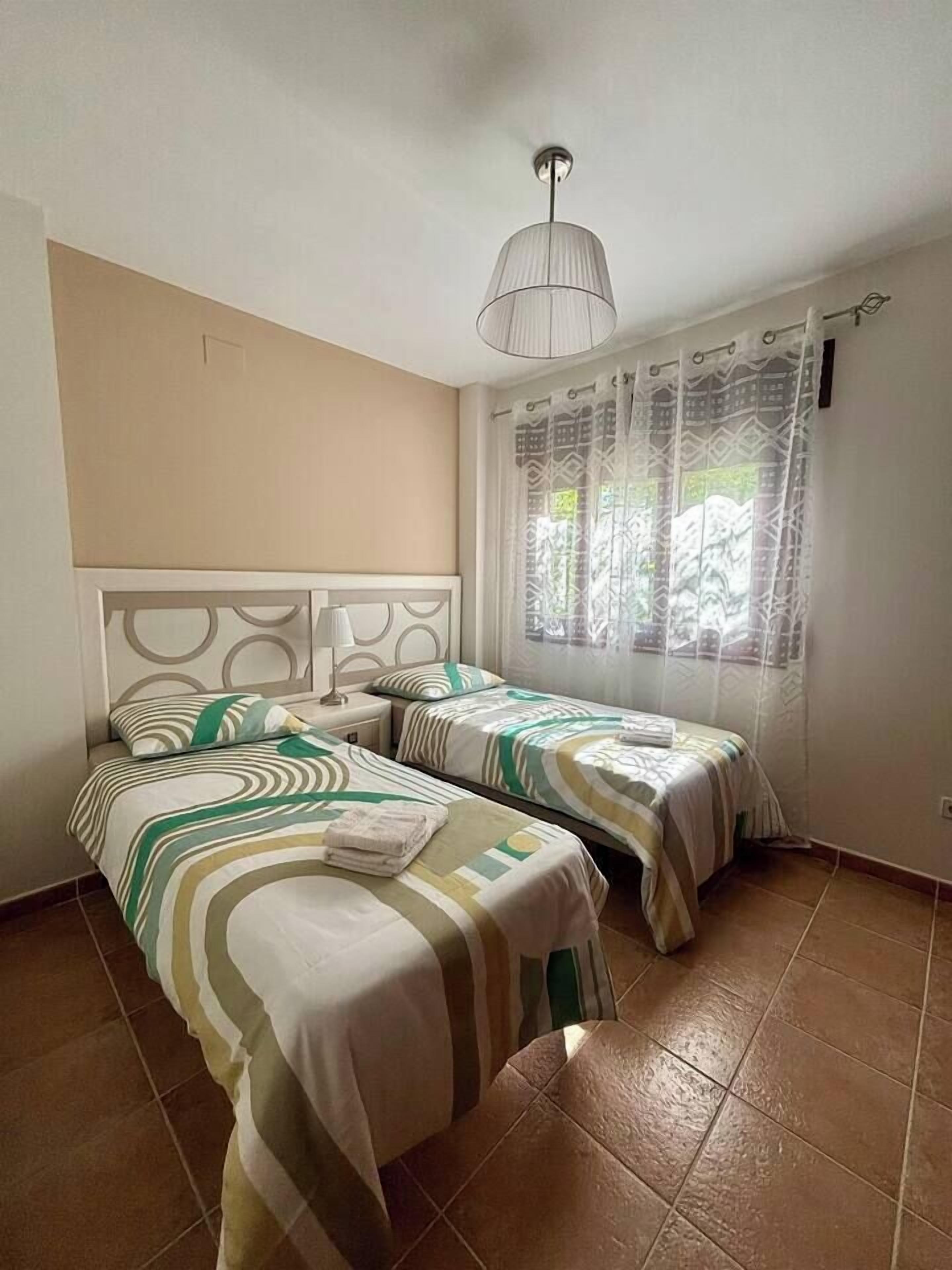 A room with two single beds, a ceiling light, a window with patterned curtains, and a tiled floor.