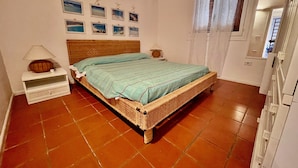 A bedroom with a wicker bed, a nightstand with a lamp, a wall with framed pictures, and a window with curtains. - Casa Petralana by Wonderful Italy (Olbia)