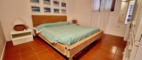 Un dormitorio con una cama de mimbre, una mesita de noche con lámpara, una pared con cuadros enmarcados y una ventana con cortinas.