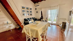 A dining area with a table set for four, a sofa, and a window with curtains. - Casa Petralana by Wonderful Italy (Olbia)