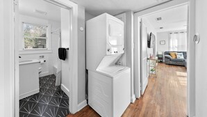 A laundry room with a washing machine and dryer, a window with blinds, and a hallway leading to a living area with a sofa. - Sunny Side Murry Hill Home (Jacksonville)