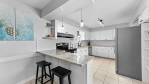 A modern kitchen with white cabinets, a granite countertop, and a stainless steel refrigerator. There are two black bar stools and pendant lights above the island. - Sunny Side Murry Hill Home (Jacksonville)
