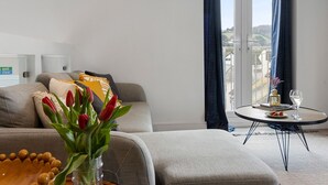 Ett modernt vardagsrum med en soffa, ett litet runt bord med ett glas vatten och en tallrik mat, och en utsikt över staden genom stora fönster.