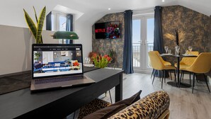 Ett modernt vardagsrum med en bärbar dator, ett matbord med gula stolar, en soffa med kuddar i leopardmönster, en växt och en tv som är monterad på väggen.