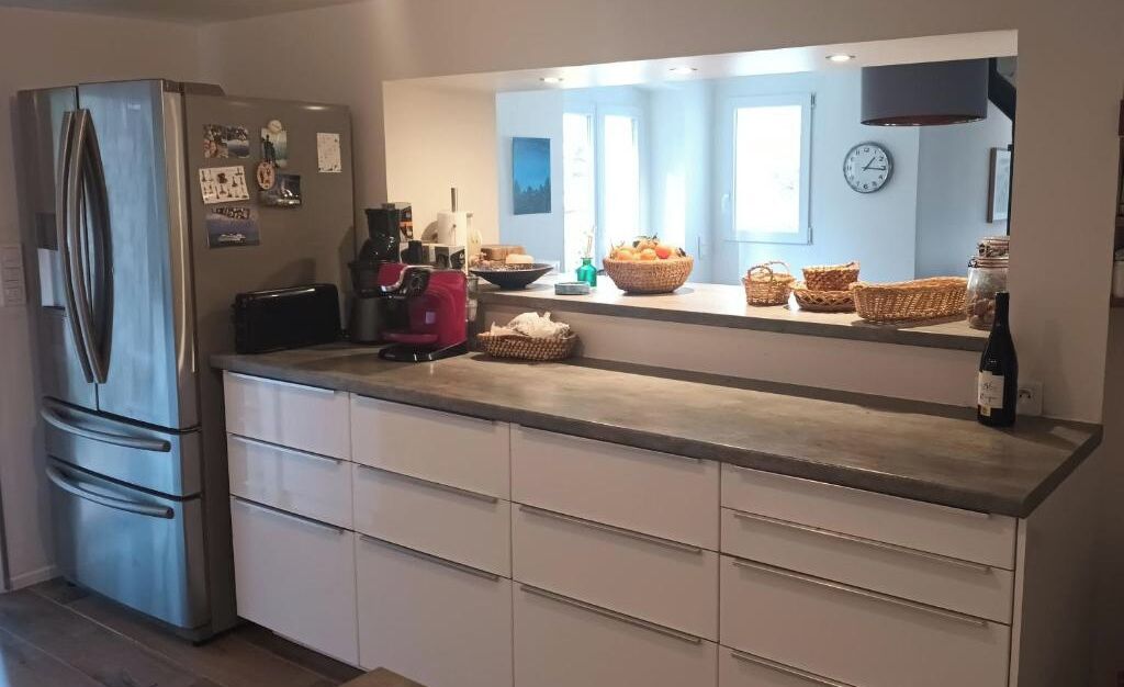 A modern kitchen with a stainless steel refrigerator, white cabinetry, a built-in oven, and a large window providing natural light.