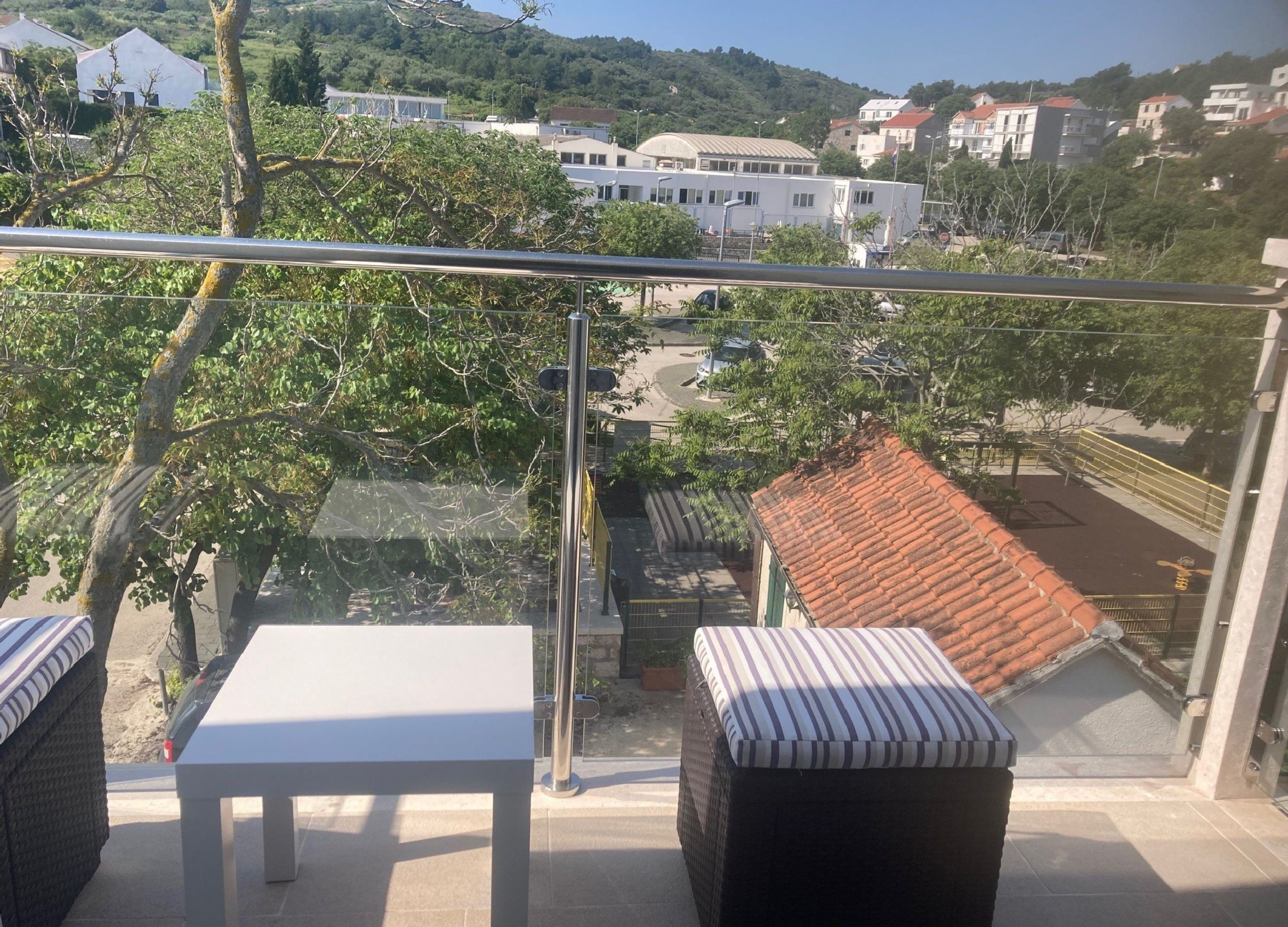 A balcony with a white table and a striped cushion, overlooking a residential area with houses and a hilly landscape.
