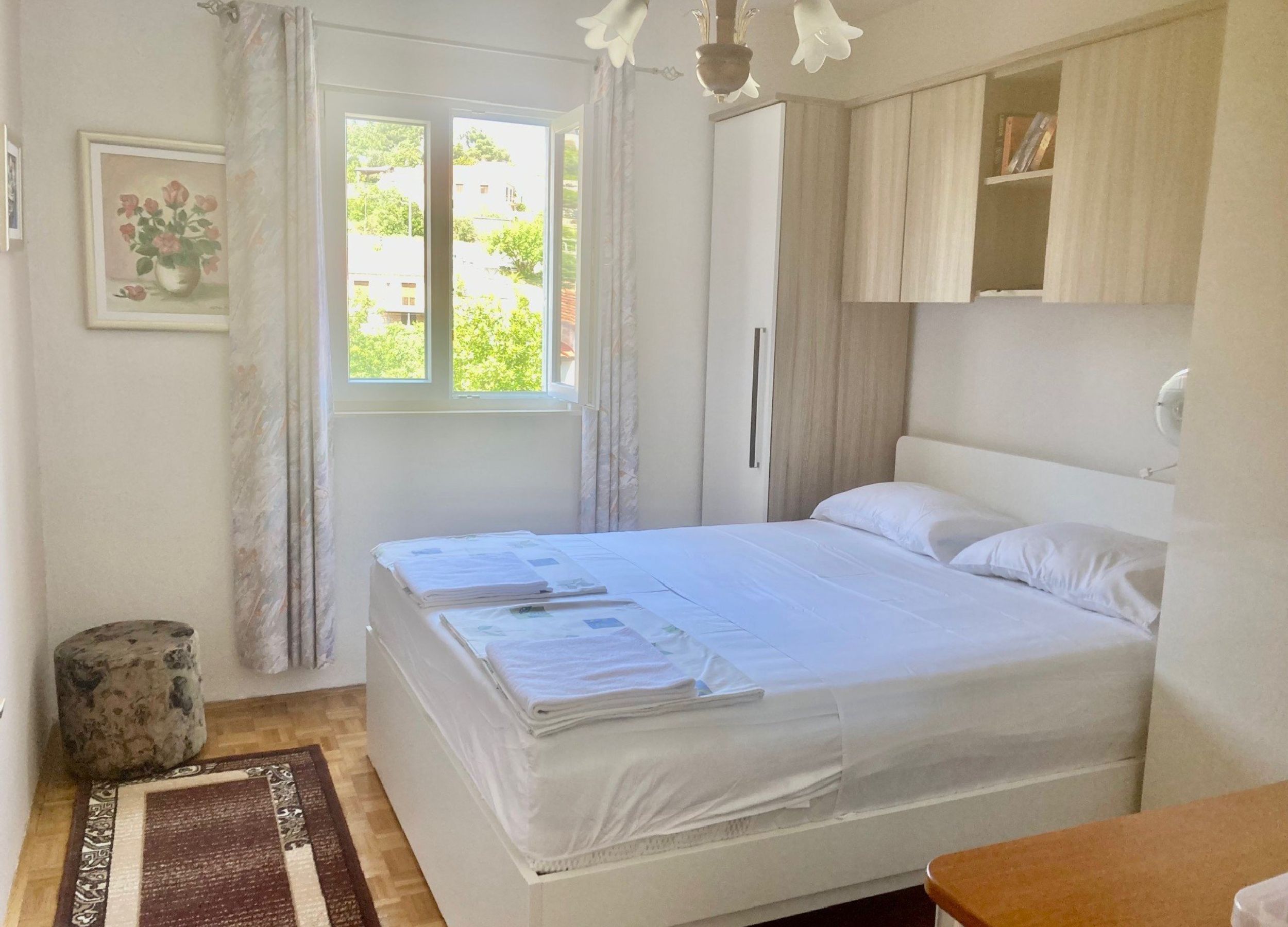 A bedroom with a bed, a window with curtains, a bedside table, a lamp, and a small stool.