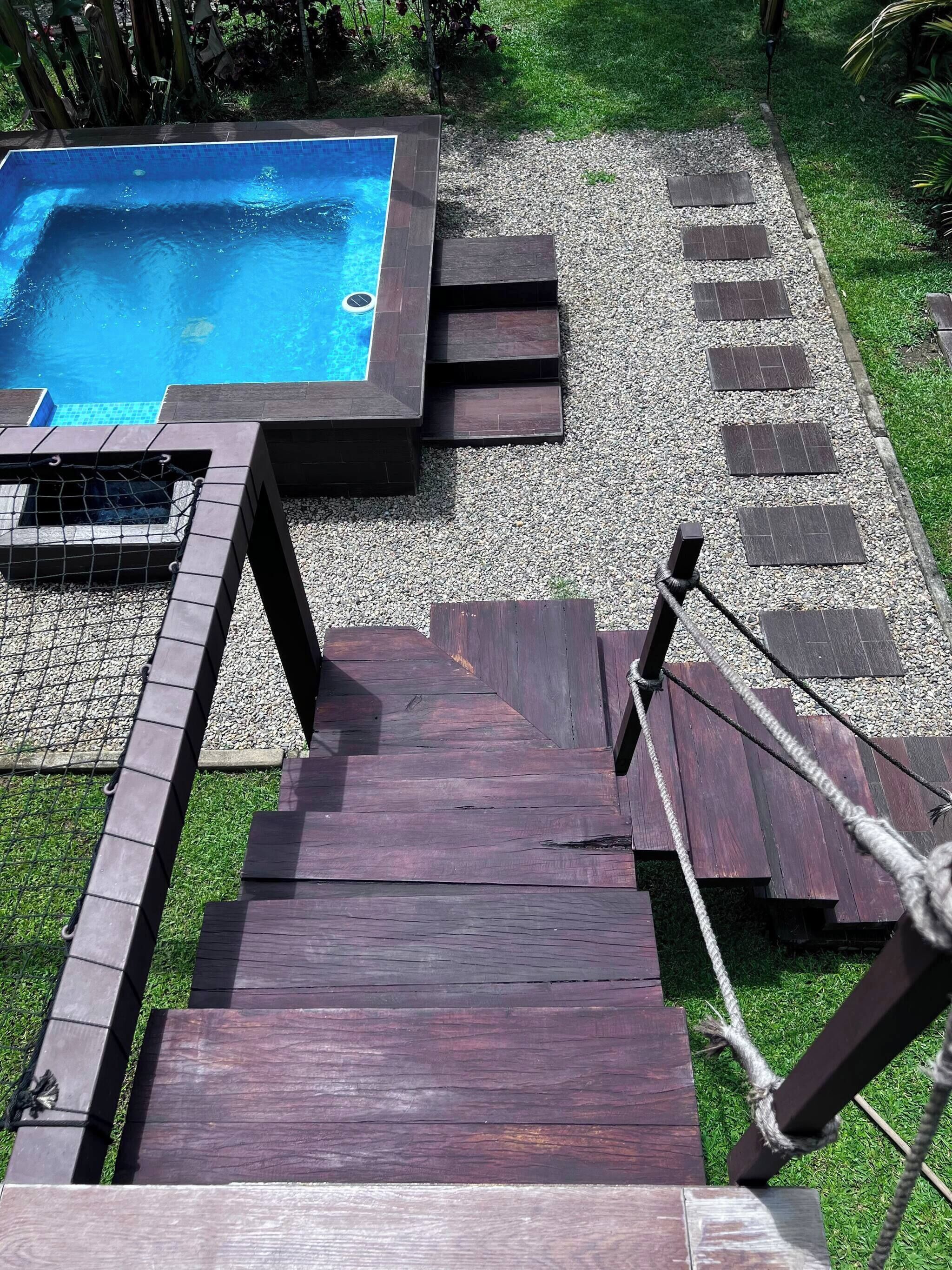 Een houten trap leidt naar een zwembad met een jacuzzi, omringd door een grindpad en groen.