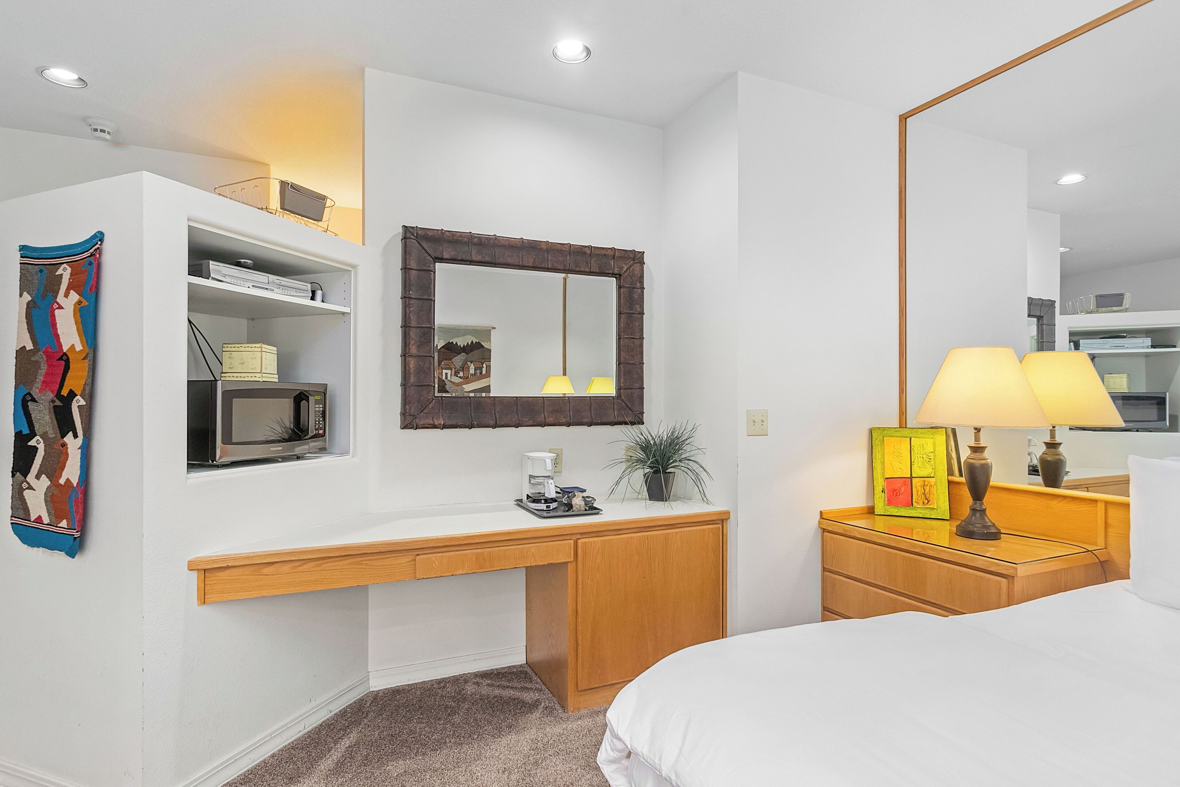 A hotel room with a white desk, a microwave, a coffee maker, a lamp, and a bed.