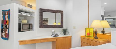A hotel room with a white desk, a microwave, a coffee maker, a lamp, and a bed.