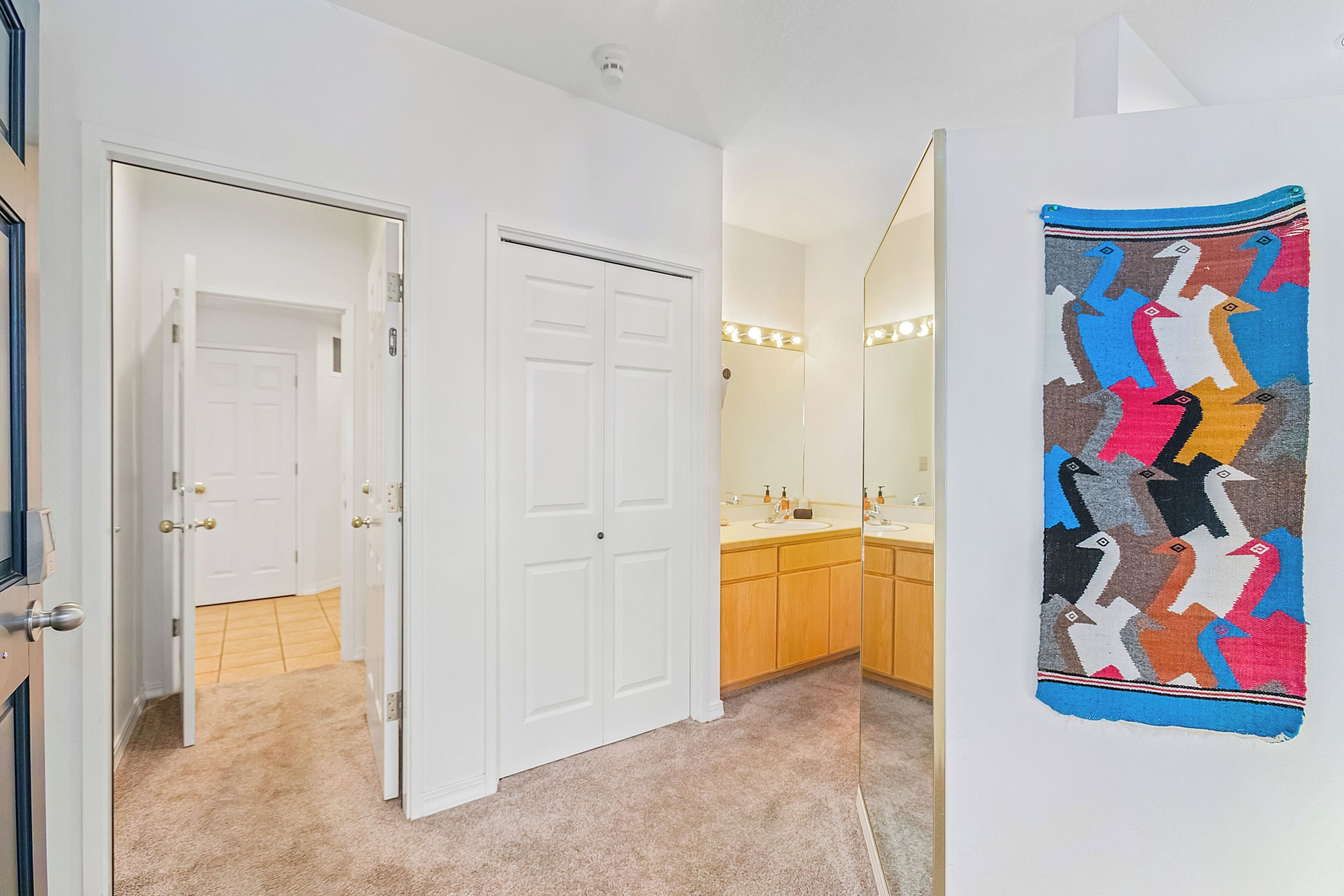 A hallway with a white door, a beige carpet, and a bathroom with wooden cabinets and a mirror.