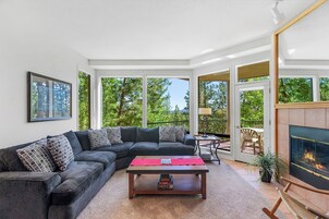 Una sala de estar moderna con un sofá modular, una mesa de centro y una chimenea. Hay un balcón con mobiliario exterior y vistas a árboles.