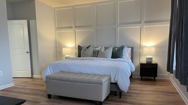 A bedroom with a bed, an ottoman, two bedside tables, a door, and a wall with paneling.
