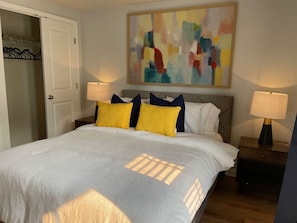 A bedroom with a bed, two bedside tables, a nightstand, a door, and a colorful abstract painting on the wall.