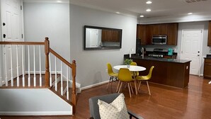 A spacious living area with a wooden staircase, a dining table with yellow chairs, and a kitchen area with wooden cabinets and a microwave.