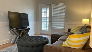 A living room with a brown leather sofa, a black ottoman, a TV on a stand, and a door with blinds.