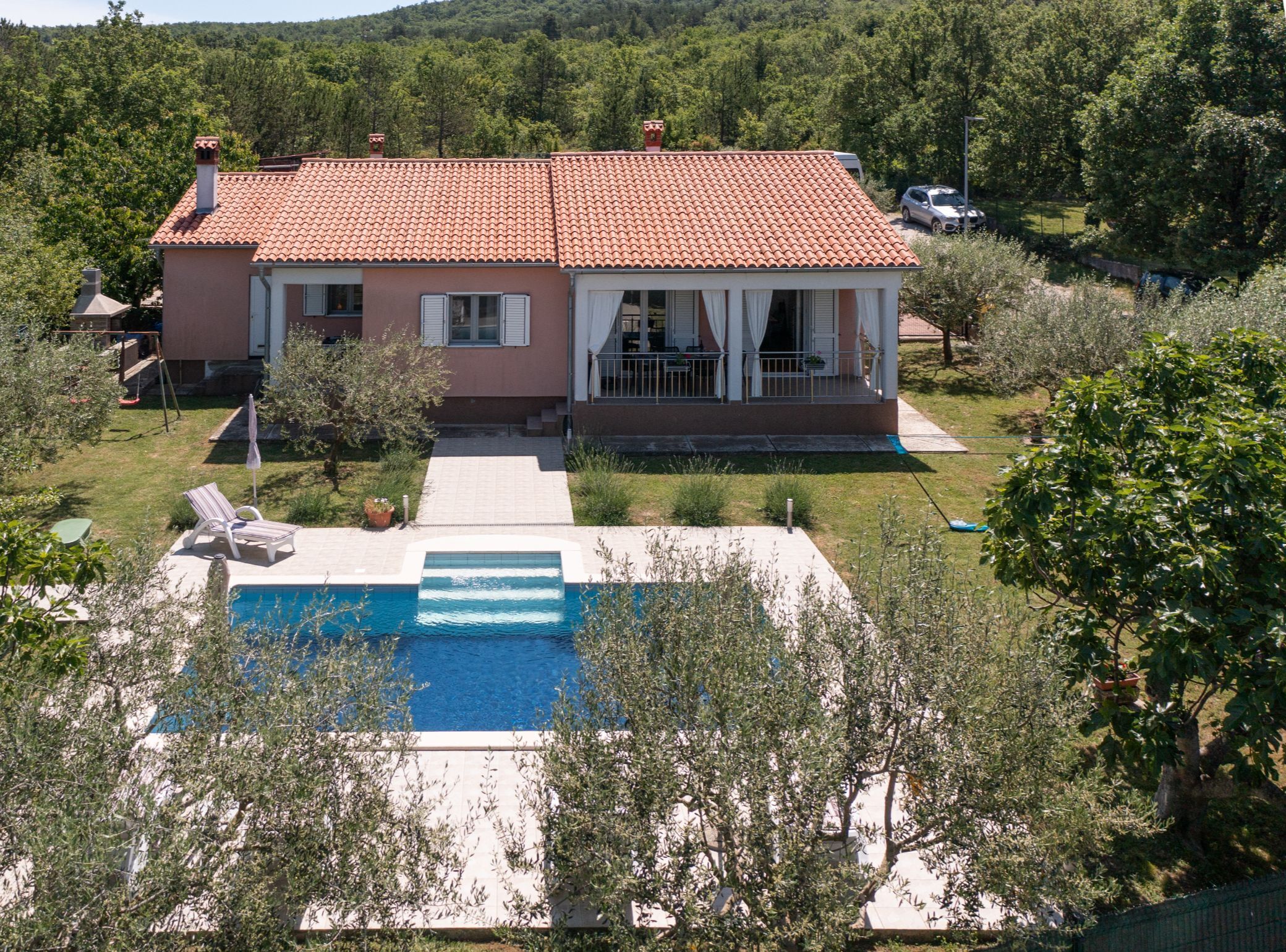 A two-story house with a swimming pool, surrounded by greenery and hills.