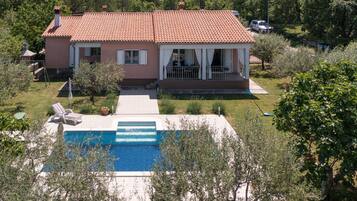 A two-story house with a swimming pool, surrounded by greenery and hills.