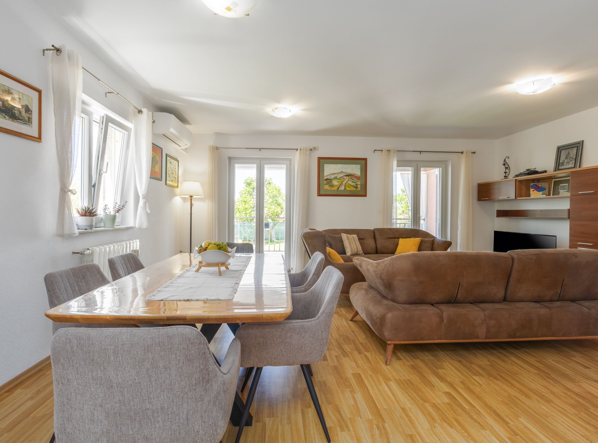A modern living room with a wooden dining table, grey chairs, a brown sofa, and a TV.
