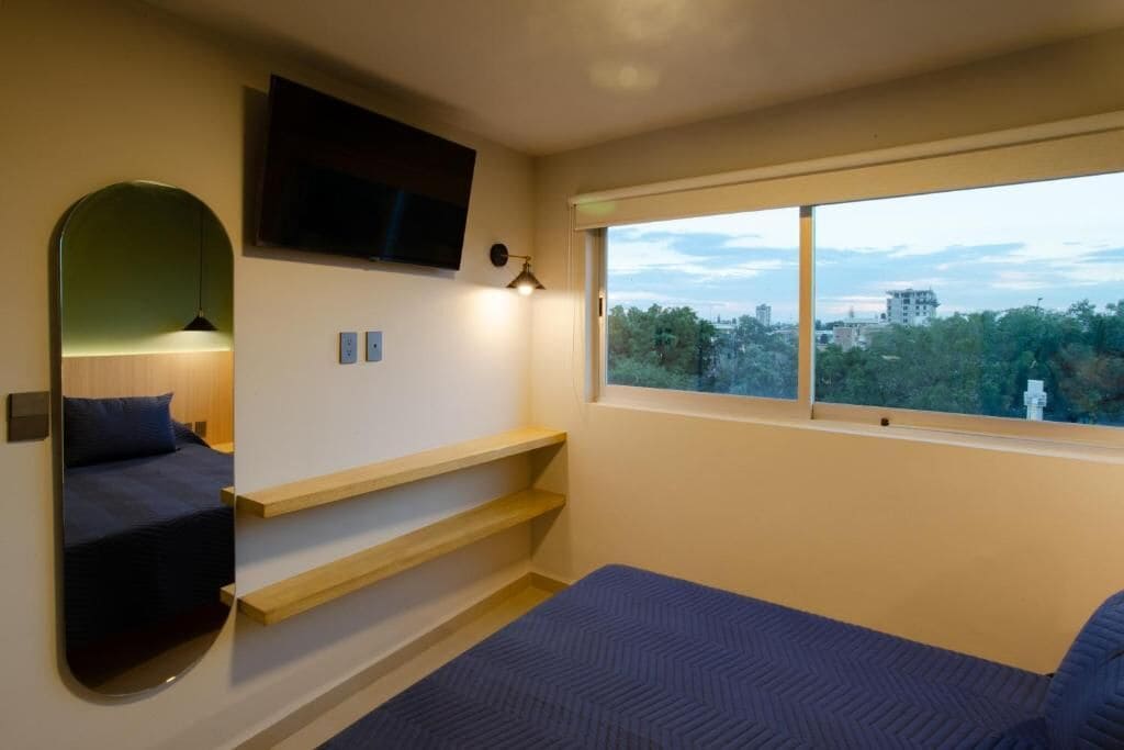 Un dormitorio con cama, televisor montado en la pared, ventana con vista a la ciudad y estante con espejo.