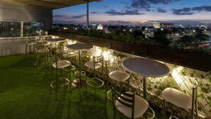 An outdoor terrace with round tables and chairs, surrounded by a green artificial grass area, overlooking a cityscape at dusk. - La Hospedería León (León)