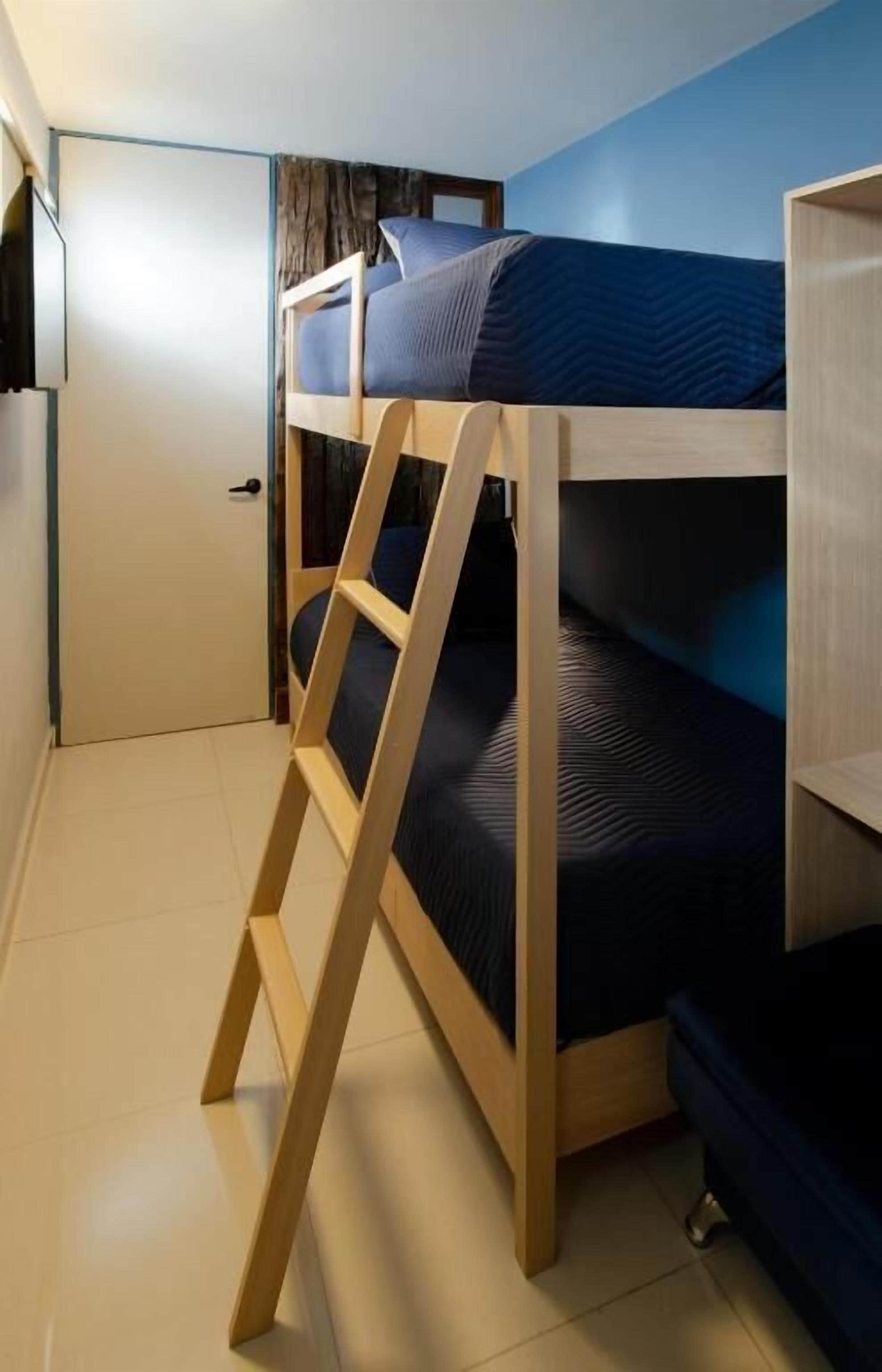 Una cama litera con escalera de madera, colchón azul oscuro y una puerta blanca al fondo.