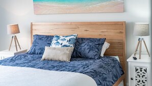 A bedroom with a wooden headboard, a bed with blue floral bedding, two nightstands with lamps, and a beach-themed painting above the bed.