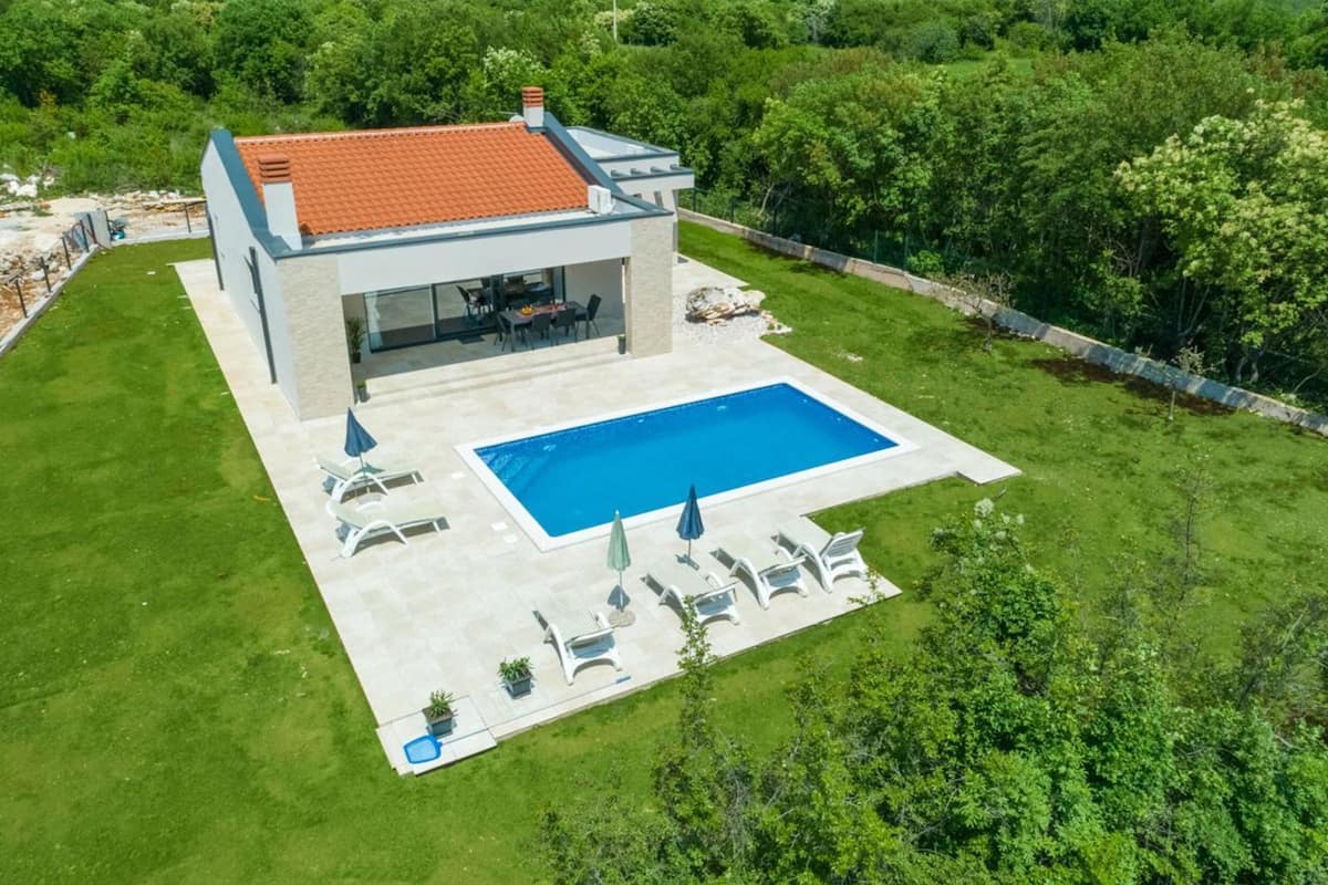 A modern house with a swimming pool, a patio with lounge chairs, and a view of the surrounding greenery.