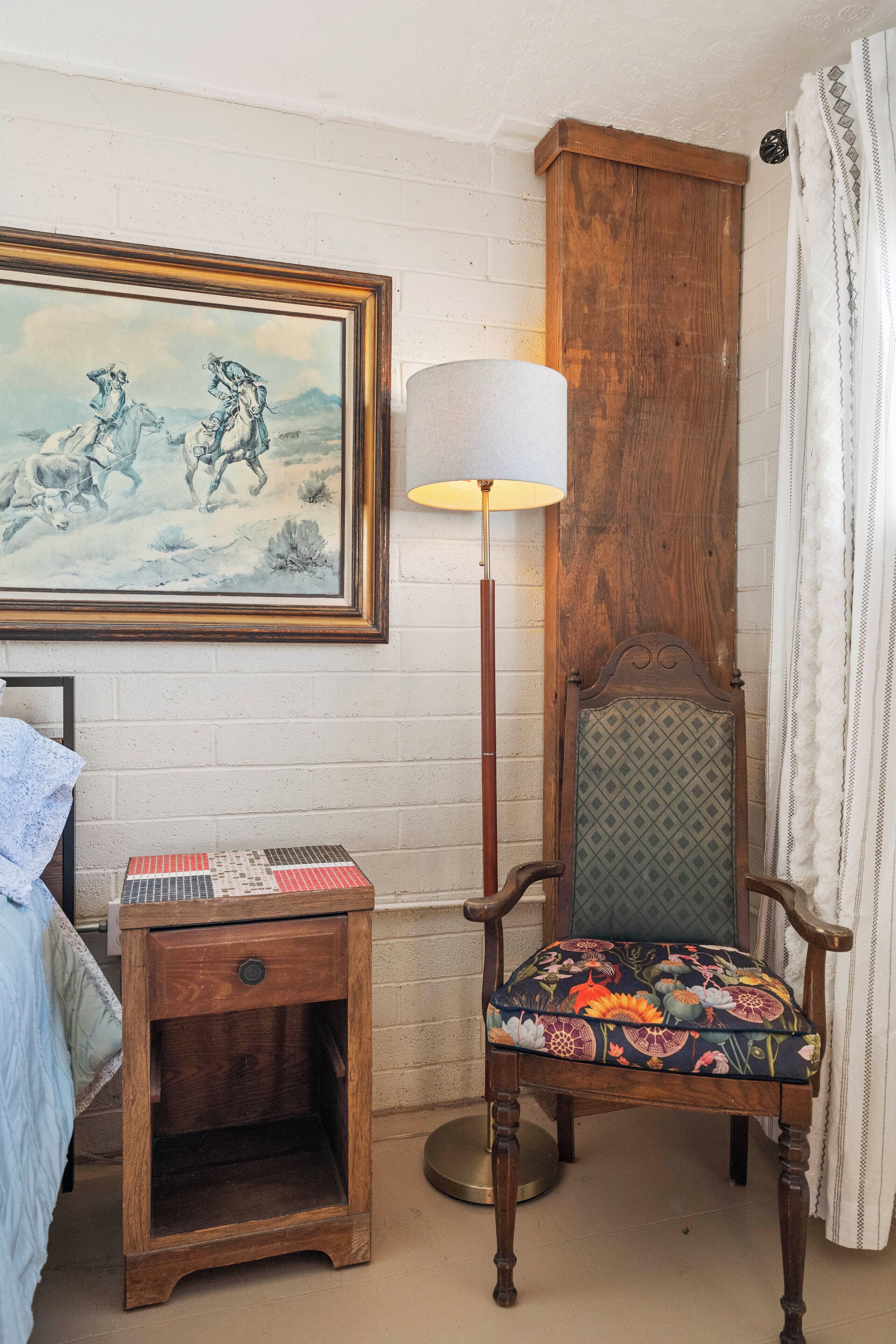 A room with a wooden nightstand, a chair with floral upholstery, a floor lamp, and a framed painting on the wall.