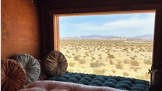 Un dormitorio con un ventanal grande, una cama con edredón verde y rosa, y dos almohadones decorativos.
