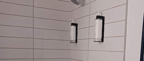 A shower with a handheld showerhead, a wall-mounted faucet, and two bottles of shampoo on a black holder.