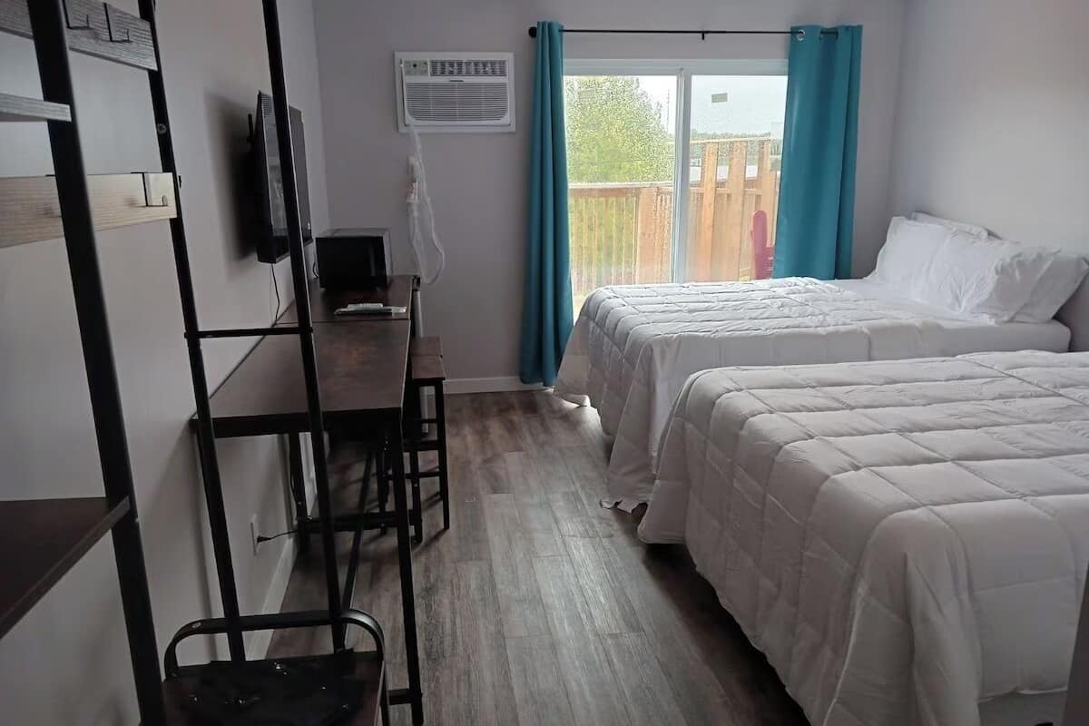 A bedroom with two beds, a desk, and a chair. There is a window with curtains and a view of the outside.