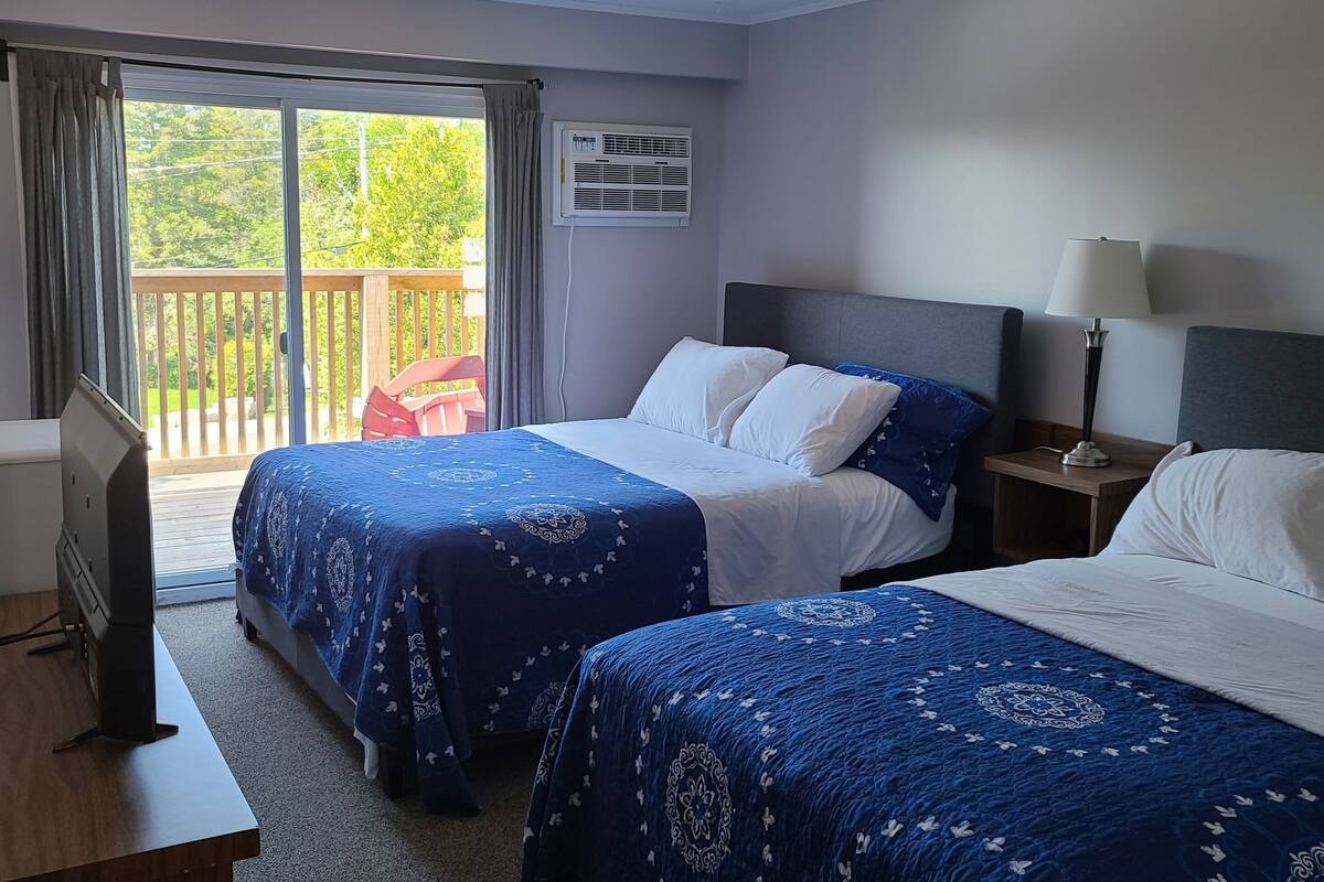A hotel room with two beds, a TV, a desk, and a balcony with a view of greenery.