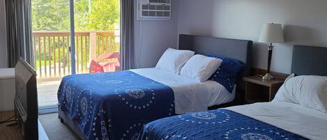 A hotel room with two beds, a TV, a desk, and a balcony with a view of greenery.