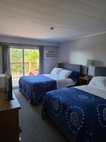 A hotel room with two beds, a TV, a desk, and a balcony with a view of greenery.