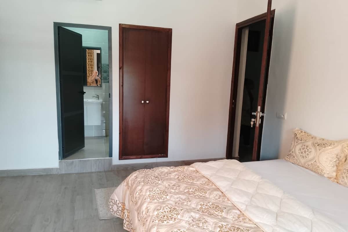 A bedroom with a bed, a wooden wardrobe, and a bathroom visible through an open door.