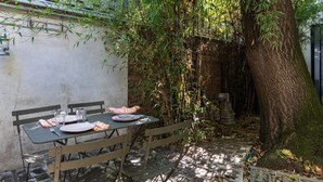 An outdoor dining area with a table set for two, surrounded by greenery and a tree.