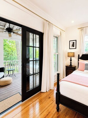 A bedroom with a large sliding glass door leading to a balcony, a bed with a white and pink bedding, a nightstand with a lamp, and a framed picture on the wall.