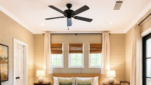 A bedroom with a large bed, a ceiling fan, three windows with blinds, a bedside table with a lamp, and a chair.