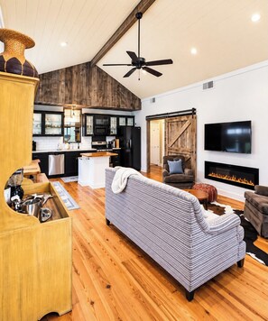 A modern living room with a fireplace, a sofa, a television, and a kitchen area with wooden cabinets. - The NestEgg, Firepit+Pool, VelvetDitchVillas (Oxford)