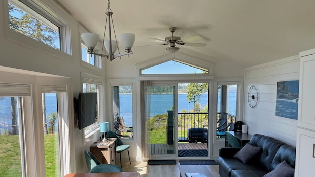 En rummelig opholdsstue med et træbord, en sort lædersofa og et fjernsyn monteret på væggen. Der er en skydedør med glas, som fører til en terrasse med udsigt over vandet.