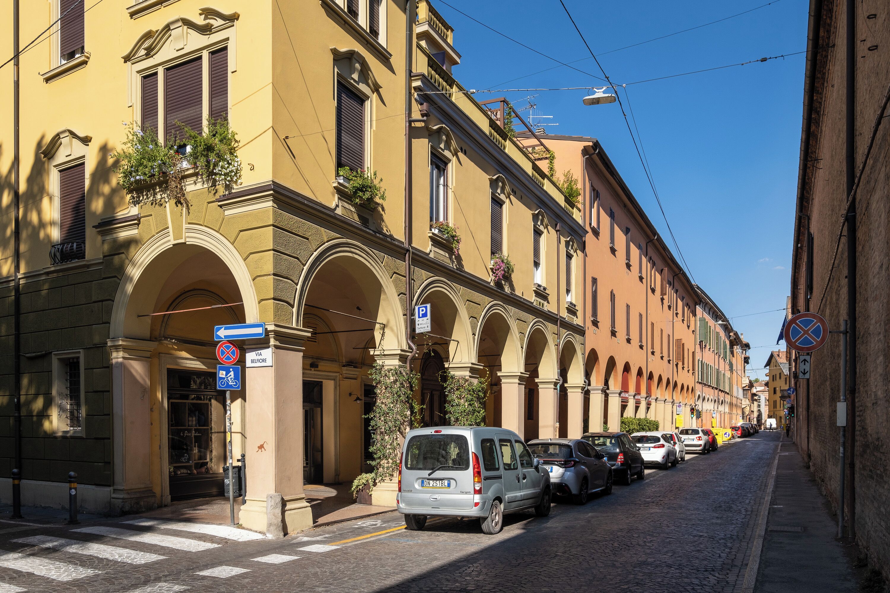 A street with parked cars, a building with arches, and a clear blue sky.